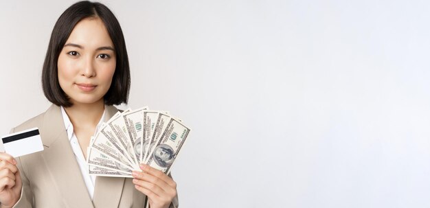 Zbliżenie azjatyckiej bizneswoman biurowej pani pokazującej kartę kredytową i pieniądze w dolarach stojących w garniturze na białym tle