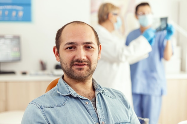 Zbliżenie cierpliwego mężczyzny patrzącego w kamerę, czekając na lekarzy, aby rozpocząć chirurgię stomatologiczną, siedząc na fotelu dentystycznym