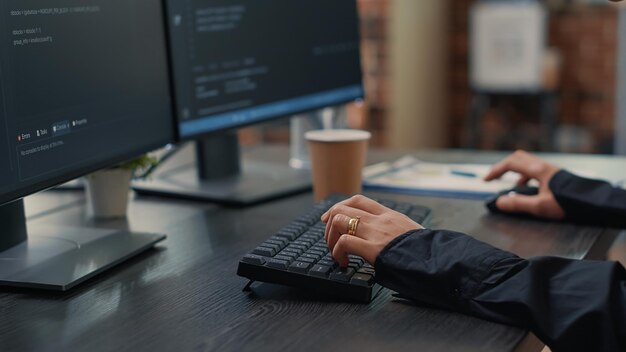 Zbliżenie dłoni dewelopera wpisując kod na klawiaturze, patrząc na ekrany komputerów z interfejsem programowania. Programista siedzący przy biurku z algorytmem pisania schowka.