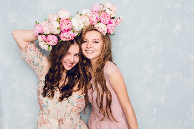 Zbliżenie dwóch pięknych dziewczyn stojących w pracowni, które bawią się głupio z diademami kwiatów na głowach.