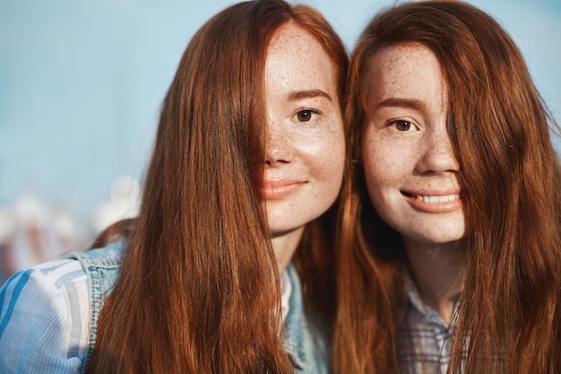 Zbliżenie dwóch rudowłosych sióstr z uroczymi piegami i uroczym uśmiechem dotykającym twarzy i szeroko uśmiechającym się do kamery zakrywającej połowę twarzy włosami wyglądającymi jak bliźniaczki.