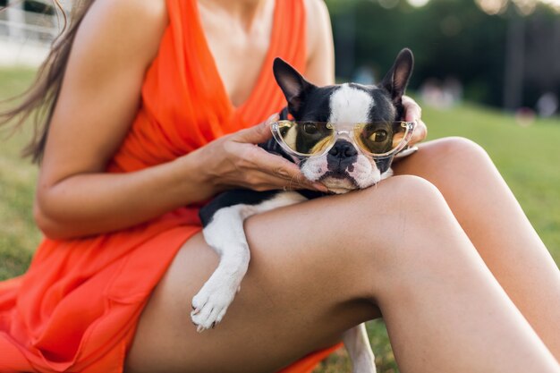 Zbliżenie kobiety siedzącej na trawie w letnim parku trzymającej boston terrier, zabawny pies w stylowych okularach przeciwsłonecznych, dziewczyna bawiąca się zwierzakiem, dobra zabawa