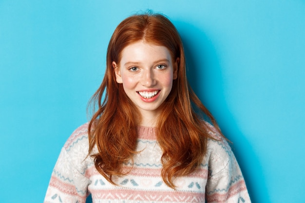 Zbliżenie ładny rudzielec dziewczyna w swetrze uśmiechający się szczęśliwy w aparacie, stojąc na niebieskim tle.