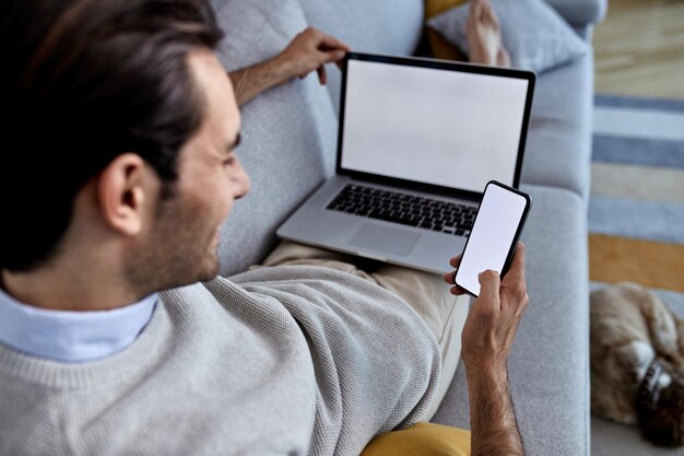 Zbliżenie mężczyzny wypoczywającego na kanapie i używającego smartfona podczas pracy na laptopie