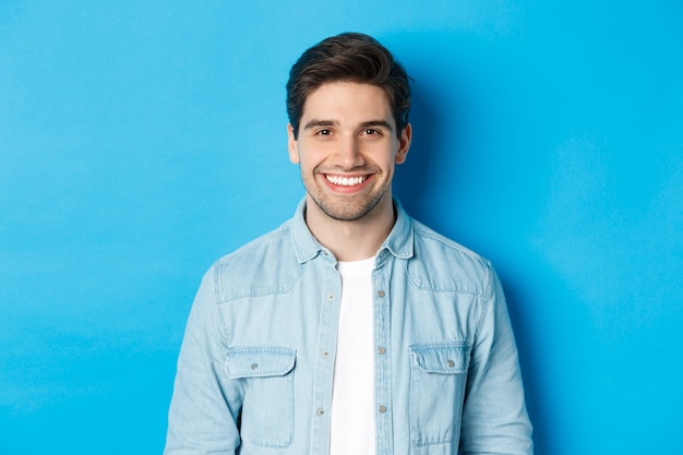 Zbliżenie młodego człowieka sukcesu uśmiechającego się do kamery, stojącego w swobodnym stroju na niebieskim tle