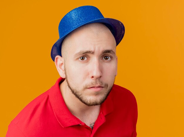 Zbliżenie młodego przystojnego faceta słowiańskich partii na sobie kapelusz partii patrząc z przodu na białym tle na pomarańczowej ścianie z miejsca na kopię