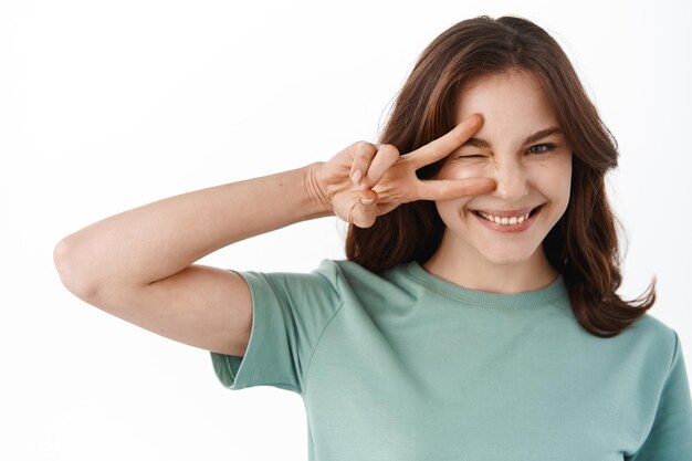 Zbliżenie młodej pozytywnej kobiety mruga do ciebie i uśmiecha się, pokazując gest pokoju w kształcie litery V w pobliżu oka, stojąc w letnim stroju na białym tle