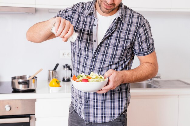 Zbliżenie: młody człowiek, dodając sól do sałatki warzywnej podczas gotowania w kuchni