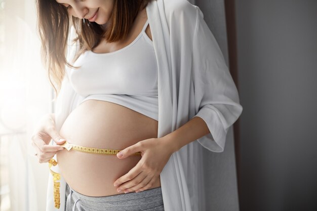 Zbliżenie na ciężarną kobietę mierzącą jej brzuch, aby śledzić rozwój jej dziecka. Szczęśliwa mama dbająca o zdrową ciążę.