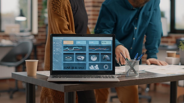 Zbliżenie na nowoczesny laptop z wykresami stawek na wyświetlaczu, podczas gdy mężczyzna i kobieta pracują nad projektem biznesowym. Ekran komputera z informacjami o wykresie danych i analizą finansów na biurku.