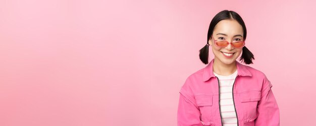 Zbliżenie na piękną azjatycką modelkę w stylowych okularach przeciwsłonecznych pozujących na różowym tle w modnej przestrzeni kopii stroju