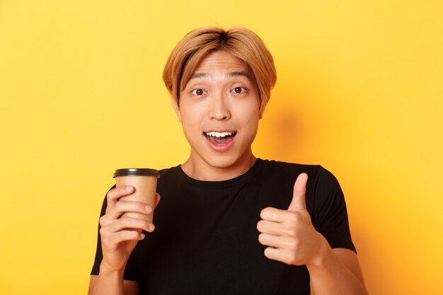 Zbliżenie na zaskoczonego przystojnego Azjatę polecającego kawiarnię, trzymającego filiżankę kawy i pokazującego z aprobatą kciuki, uśmiechającego się zadowolony nad żółtą ścianą.