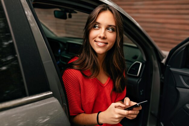 Zbliżenie na zewnątrz portret uroczej słodkiej dziewczyny z długimi falującymi włosami i cudownym uśmiechem przewija smartfona siedząc w samochodzie