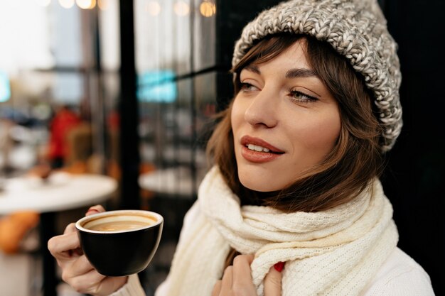Zbliżenie na zewnątrz zdjęcia europejskiej uroczej kobiety noszącej czapkę z dzianiny i szalik pijący kawę w kawiarni miejskiej ze światłami świątecznego nastroju
