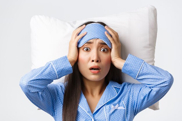 Zbliżenie paniki azjatyckiej słodkiej dziewczyny leżącej w łóżku na poduszce w masce do spania i piżamie, wyglądające na zaniepokojone i zaniepokojone, zdaje sobie sprawę, że stało się coś złego, pozują zmartwione białe tło