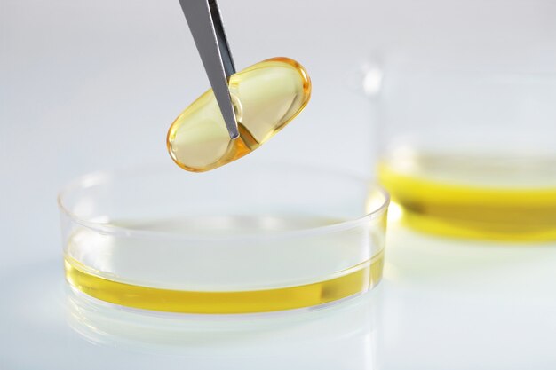 Zbliżenie pęsety trzymającej przezroczystą żółtą kapsułkę nad żółtym płynem w szklanym naczyniu w laboratorium