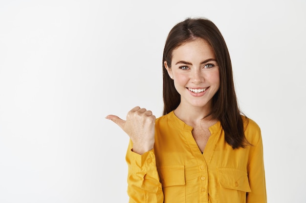 Zbliżenie: pewna siebie i szczęśliwa brunetka, wskazując kciukiem w lewo, sprawdzając ofertę promocyjną na białej przestrzeni kopii.