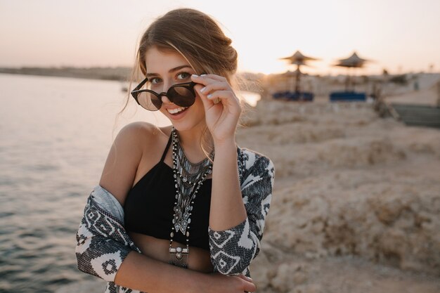Zbliżenie portret atrakcyjna młoda dziewczyna zdejmując stylowe okulary przeciwsłoneczne, na zachód słońca, na plaży z zmysłowym wyglądem. Ubrana w modny czarny top, naszyjnik, kardigan z ozdobami. Ośrodek wypoczynkowy, wczasy, wakacje.