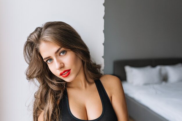 Zbliżenie portret atrakcyjna urocza dziewczyna z długimi brunetkami w nowoczesnym mieszkaniu. Seksowna koszulka, prawdziwe emocje, czerwone usta. Poranek, modny model, relaks.