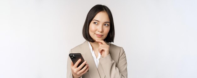Zbliżenie portret koreańskiej kobiety korporacyjnej damy w garniturze za pomocą telefonu komórkowego i uśmiechniętego trzymającego smartfona stojącego na białym tle
