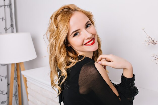 Zbliżenie portret ładna dziewczyna z blond włosy falowane, uśmiechając się, pozowanie w białym nowoczesnym pokoju. Widok z boku. Ubrana w stylową czarną sukienkę, bluzkę.