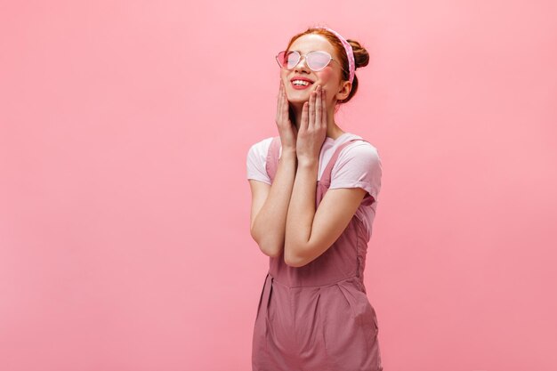 Zbliżenie portret młodej damy z opaską i różowymi okularami Kobieta w kombinezonie pozuje z uśmiechem na różowym tle