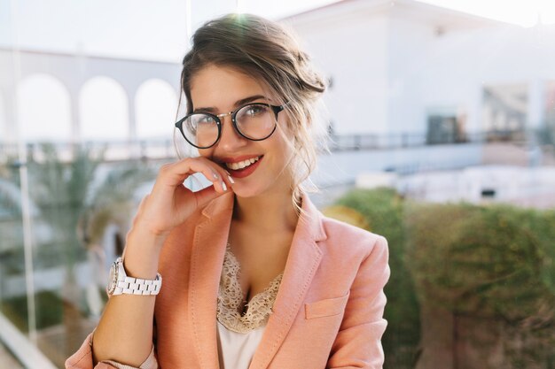 Zbliżenie portret młodej dziewczyny wspaniały w stylowych okularach, ładny student, biznes kobieta ubrana w elegancką różową kurtkę, beżową bluzkę z koronki, makijaż dzienny. Duże okno z widokiem na podwórko.