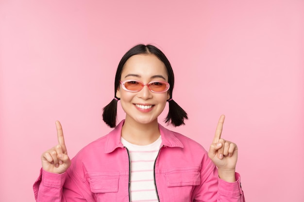 Zbliżenie portret nowoczesnej koreańskiej modelki nosi okulary przeciwsłoneczne wskazuje palce w górę pokazuje reklamę baner promocyjny różowe tło