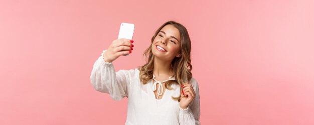 Zbliżenie portret pięknej i stylowej blond dziewczyna biorąc selfie w parku na wiosnę zebrać upodobania i zwolennicy zrobić blogger treści biorąc zdjęcie na smartfonie uśmiechający się sassy różowym tle