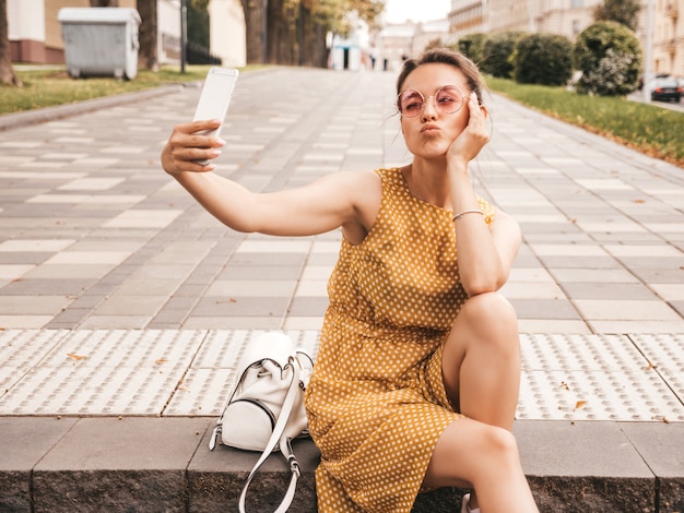 Zbliżenie Portret Pięknej Uśmiechnięte Dziewczyny Brunetka W Lato Hipster żółta Sukienka. Model Robienia Selfie Na Smartfonie. Kobieta Robienia Zdjęć W Ciepły Słoneczny Dzień Na Ulicy W Okulary Przeciwsłoneczne