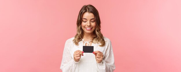 Zbliżenie portret podekscytowany i rozbawiony blond dziewczyna w białej sukni, trzymając kartę kredytową i uśmiechnięty zachwycony nie mogę oprzeć się pokusie, aby kupić coś marnować pieniądze online zakupy różowy tło