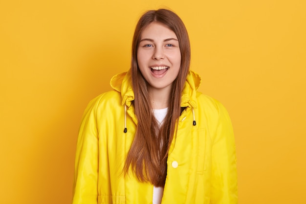 Zbliżenie portret szczęśliwa uśmiechnięta studencka dziewczyna jest ubranym kurtkę, wrzeszczy coś szczęśliwie, stać odizolowywam nad kolor żółty ścianą, wyrażający pozytywne emocje.