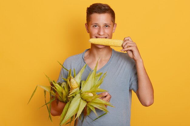 Zbliżenie portret szczęśliwy młody studencki facet gryźć kukurydzy i trzyma inne kukurudze w rękach