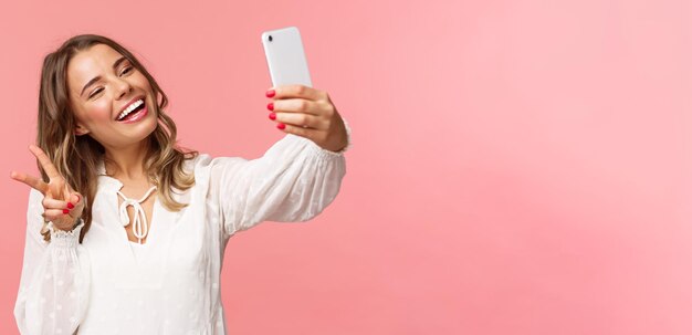 Zbliżenie portret wesoły piękny kobiecy blond dziewczyna w białej sukni biorąc selfie na telefonie komórkowym zrobić znak pokoju kawaii podczas robienia zdjęć przechwytywania chwila wiosny różowe tło