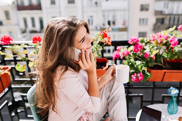 Zbliżenie portret z boku Ładna dziewczyna w piżamie jedząc śniadanie na balkonie w słoneczny poranek. Trzyma kubek, rozmawiając przez telefon z uśmiechem.