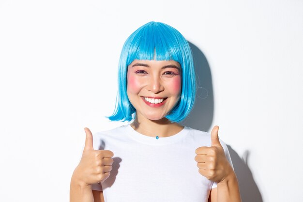 Zbliżenie: szczęśliwa azjatycka dziewczyna w niebieskiej peruce, uśmiechnięta i pokazująca kciuki w górę z aprobatą