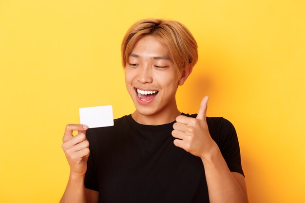 Zbliżenie szczęśliwego i zadowolonego azjatyckiego przystojnego faceta, pokazującego kartę kredytową i kciuki w górę z aprobatą, uśmiechnięty zdumiony, stojący żółtą ścianę