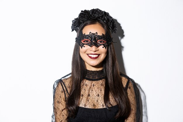 Zbliżenie: tajemnicza kobieta w gotyckim wieńcu i czarnej masce uśmiecha się do kamery, świętuje halloween, stojąc na białym tle.