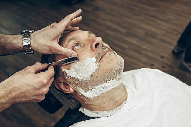 Zbliżenie z boku widok z góry przystojny starszy brodaty kaukaski mężczyzna coraz brodę pielęgnacja w nowoczesnym zakładzie fryzjerskim. Fryzjer obsługujący klienta, wykonujący strzyżenie brody za pomocą brzytwy. Koncepcja sklepu fryzjerskiego.