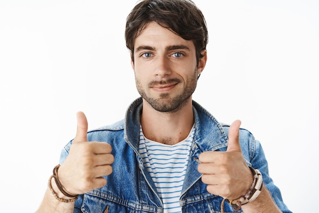 Bezpłatne zdjęcie zbliżenie zadowolonego i szczęśliwego młodego atrakcyjnego mężczyzny z niebieskimi oczami i włosiem w dżinsowej kurtce podnoszącej kciuki do góry i uśmiechającej się radośnie