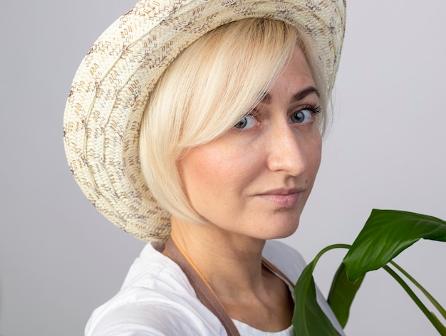 Bezpłatne zdjęcie zbliżenie zadowolonej blondynki ogrodniczki w średnim wieku w mundurze w kapeluszu stojącej za rośliną w widoku profilu