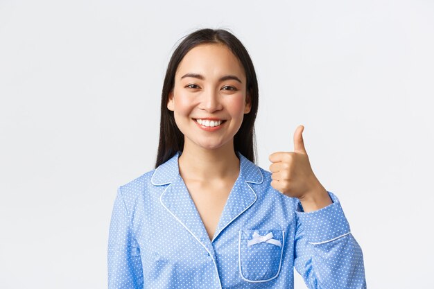 Zbliżenie zadowolonej ładnej uśmiechniętej azjatyckiej kobiety w niebieskiej piżamie pokazującej kciuki w górę w aprobacie, polecającej i gwarantującej jakość produktu, stojącej zadowolonej na białym tle