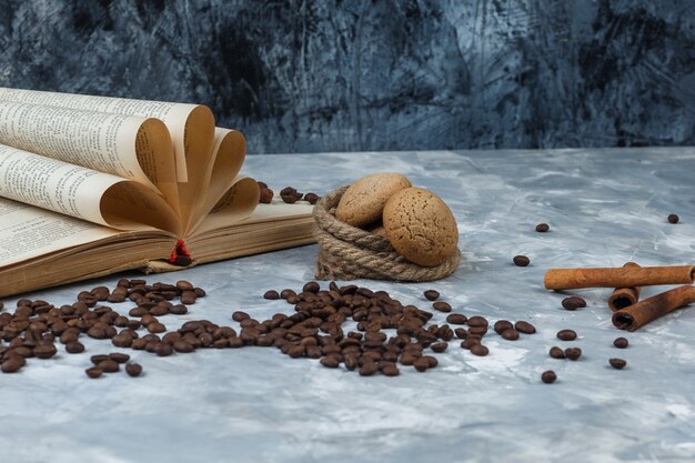 Zbliżenie ziarna kawy z książką, cynamonem, ciasteczkami, linami na ciemnym i jasnoniebieskim tle marmuru. poziomy