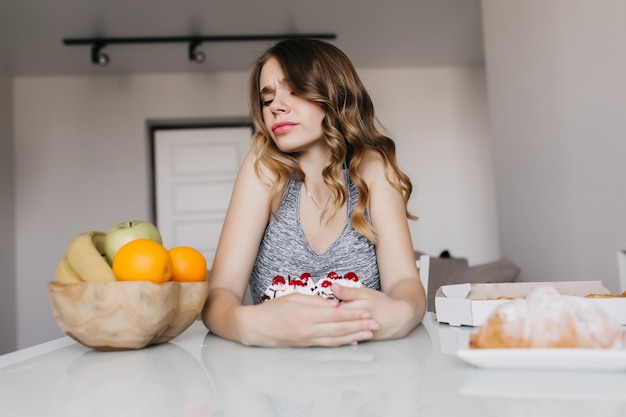 Bezpłatne zdjęcie zdenerwowana kobieta z falowanymi włosami, pozowanie podczas śniadania. ładna dziewczyna patrząc na owoce ze smutkiem i trzymając tort.