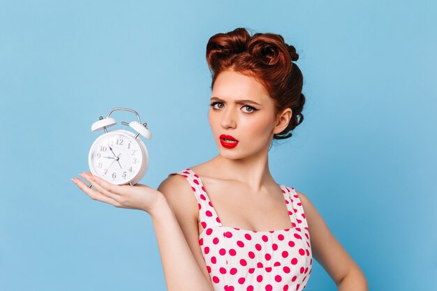 Zdenerwowana kobieta z jasnym makijażem pokazuje czas. Studio strzałów pięknej dziewczyny pinup z zegarem.