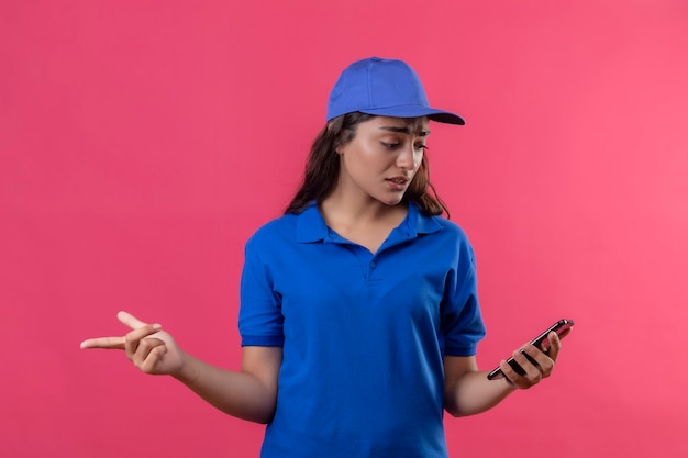 Zdenerwowana młoda dziewczyna dostawy w niebieskim mundurze i czapce, patrząc na ekran swojego smartfona ze smutnym wyrazem twarzy stojącej na różowym tle
