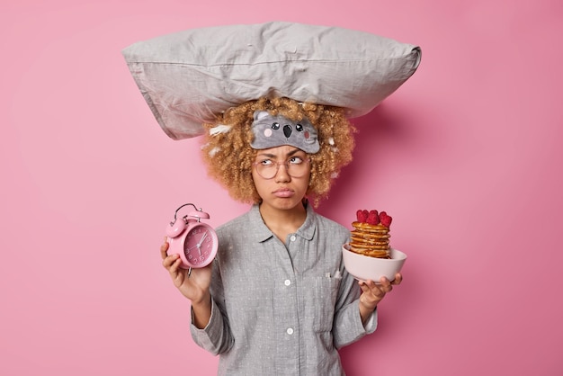 Bezpłatne zdjęcie zdenerwowana młoda kobieta ubrana w piżamę nosi poduszkę na głowie trzyma budzik i miskę naleśników z malinami ma smutne, zamyślone wyrażenie na białym tle nad różowym tle koncepcja poranka