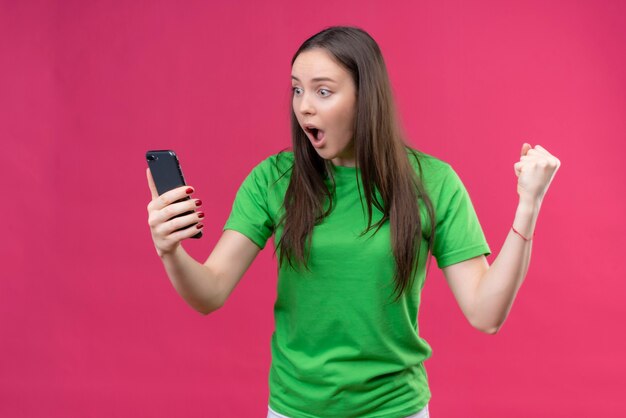 Zdenerwowana młoda piękna dziewczyna ubrana w zieloną koszulkę trzymając smartfon patrząc na ekran zdumiony i zaskoczony stojąc na na białym tle różowym tle