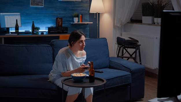Zdenerwowana, rozczarowana kobieta trzymająca joystick, grając w gry wideo w telewizji, przegrywając konkurs gier wideo online, pijąc piwo. sfrustrowana osoba grająca nad przebieraniem się w piżamę relaksującą późną nocą