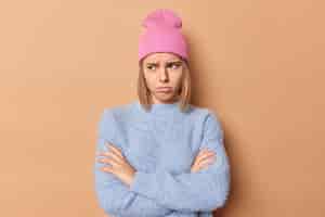 Bezpłatne zdjęcie zdenerwowana, sfrustrowana młoda kobieta z założonymi rękoma czuje się zła lub obrażona marszczy brwi twarz stoi w zamkniętej postawie odwraca wzrok nieszczęśliwie nosi różowy kapelusz i niebieski sweter na białym tle na beżowym tle.